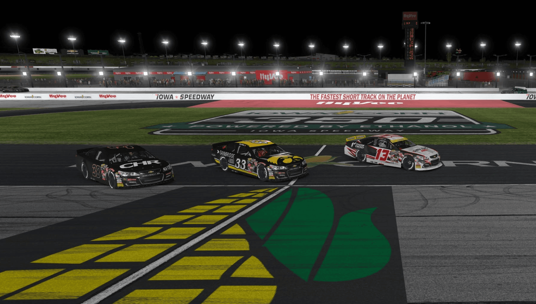 Three oval cars racing at Iowa Speedway under the lights